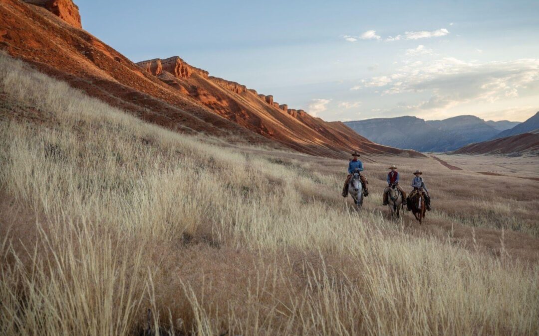 Three horseback riders crossing a grassy valley beneath red rock cliffs.