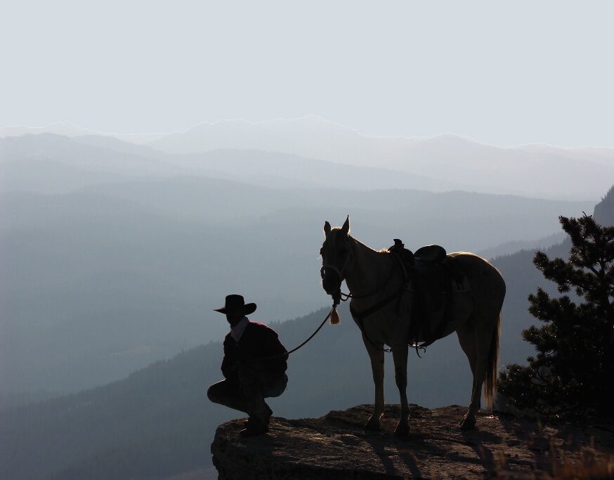 A person crouching beside a horse, silhouetted against distant mountains.