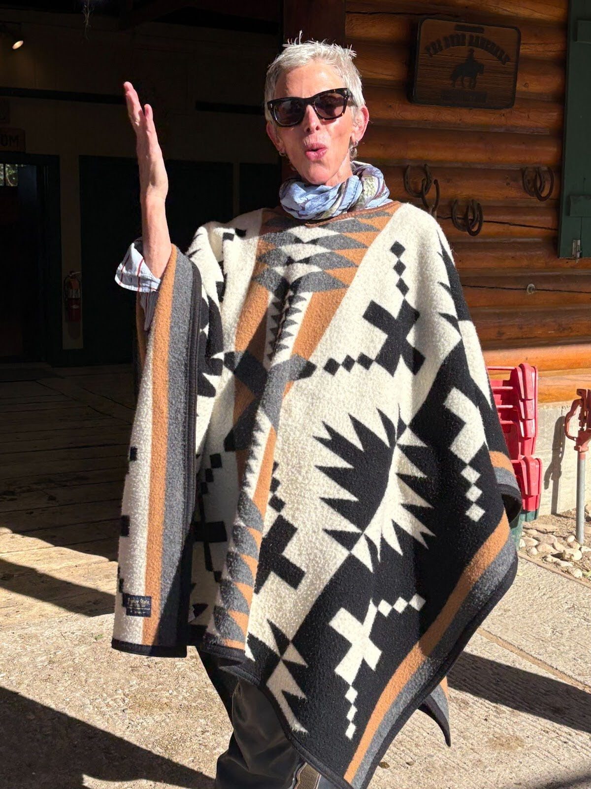 A person wearing a patterned poncho and sunglasses beside a log building.