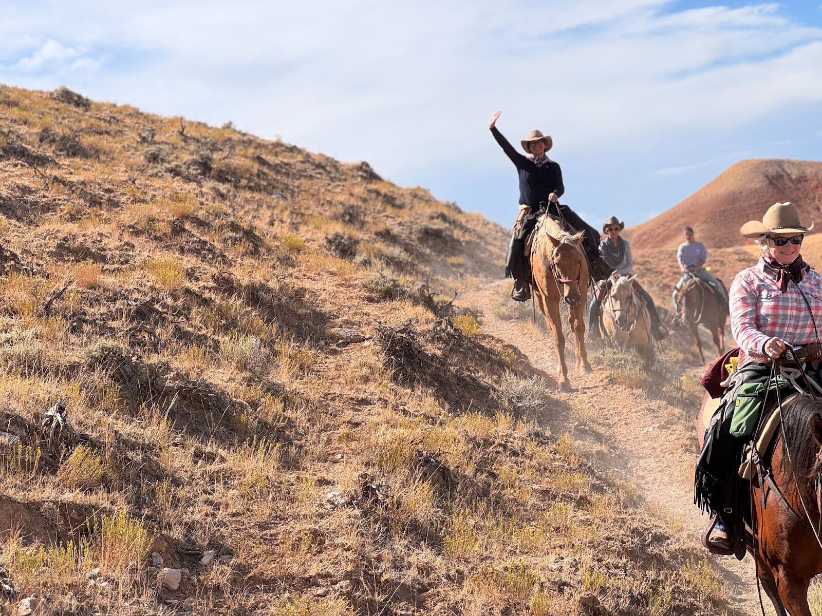 Horseback riders following a hillside trail, with one rider raising an arm.