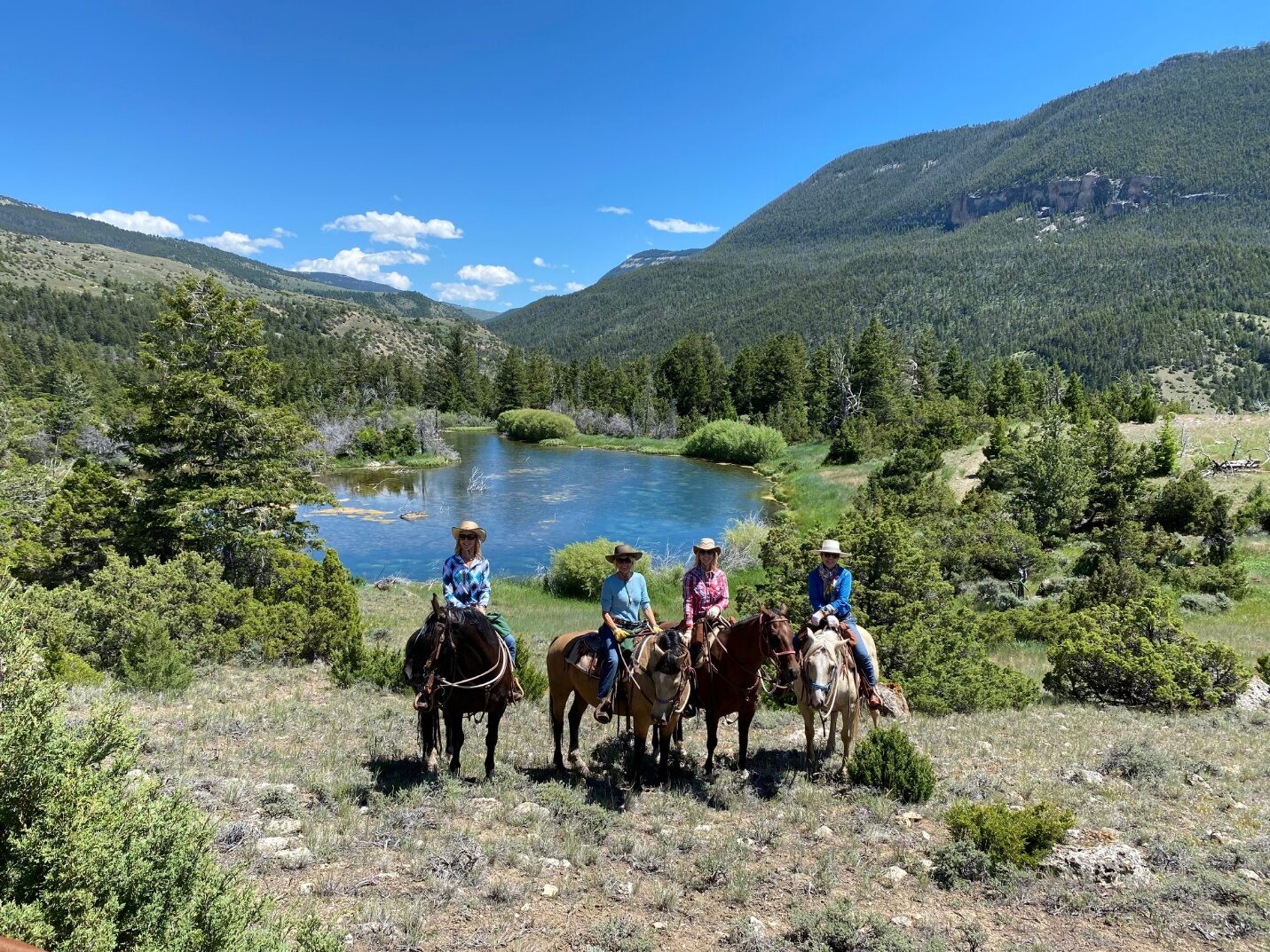 A group of horseback riders above a blue lake surrounded by forest.